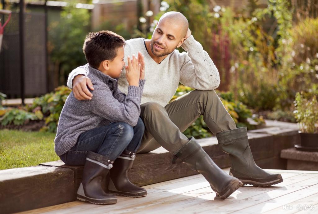 En voksen mann og en gutt sitter ved siden av hverandre ute i en hage. Mannen holder en arm om guttens skulder. Foto.