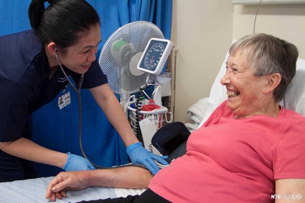 Helsefagarbeider måler blodtrykket til en kvinnelig pasient. Begge smiler. Foto.