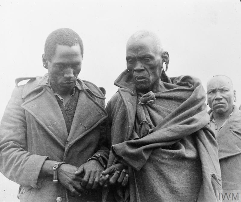 Photo: Two Black men are in the foreground of the photo. One of them is wearing metal handcuffs. The other man's wrists are obscured by his clothes. In the background, there is a third man. 