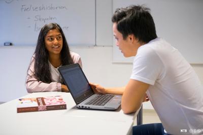 To elever sitter ved en pult med en bærbar PD og ei lærebok på. På tavla bak dem står det "Frelsehistorien" og "Skapelsen". Foto.