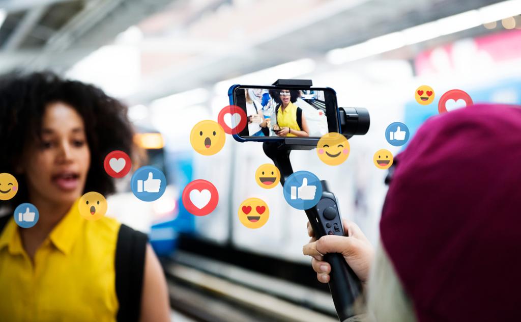 En vlogger strømmer en live video på en togplattform. Ikoner med emojier, hjerter og tomler flyr rundt henne. Foto.