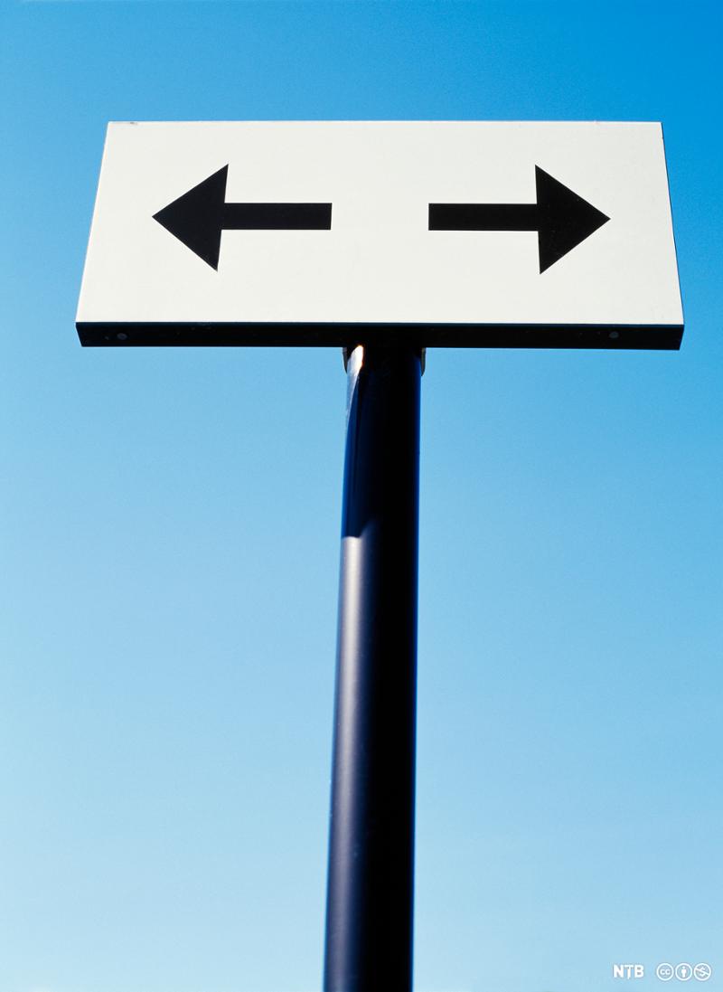 Et skilt viser en svart pil til venstre og en svart pil til høyre på hvit bakgrunn. Bak skiltet er det blå himmel. Foto.