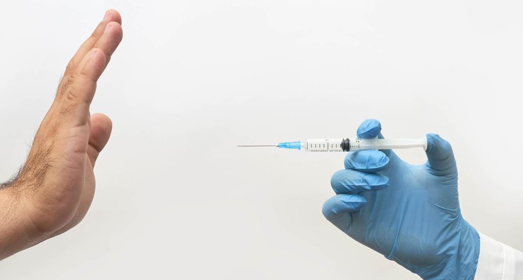 Two hands are showing: one is pointing a syringe towards the other hand. The other hand is declining the vaccine. Photo.