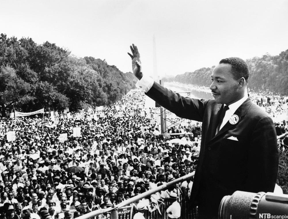 Photo: Martin Luther King Jr. at the March on Washington. We see Dr. King standing above huge crowd of people. In the background, we see the Washington Monument. 