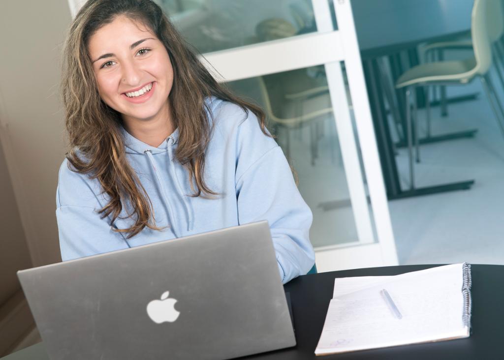 Smilende jente sitter med åpen datamaskin foran seg. Foto.