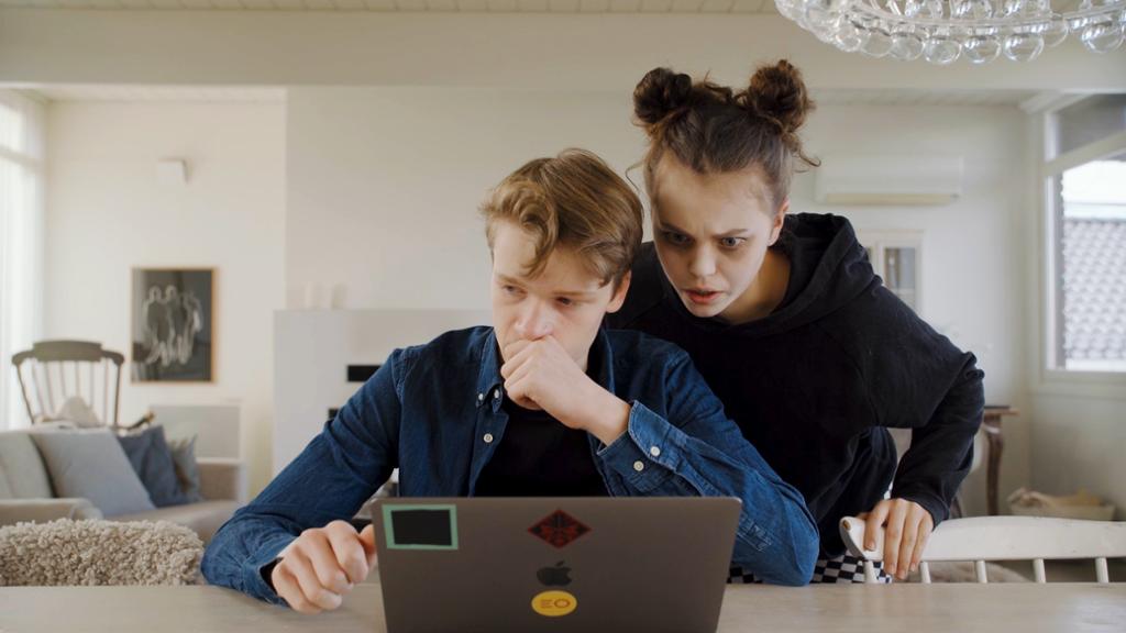 En tenåringsgutt sitter foran PC-en, mens ei jente lener seg over skulderen hans. Foto.
