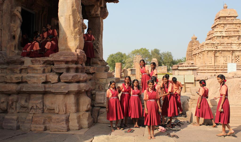 Photo: We see a two groups of Indian girls dressed in orange shirts and red dresses. They are posing in front of what looks like an ancient  temple. 