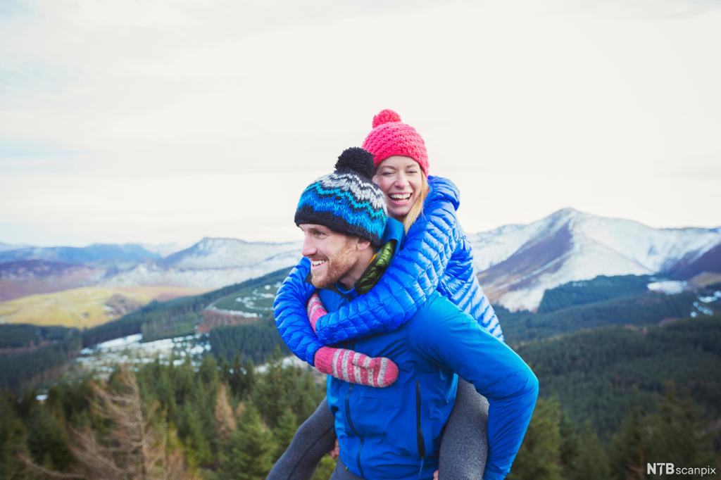 Smilende kvinne blir båret av smilende mann i fjelllandskap. Foto.