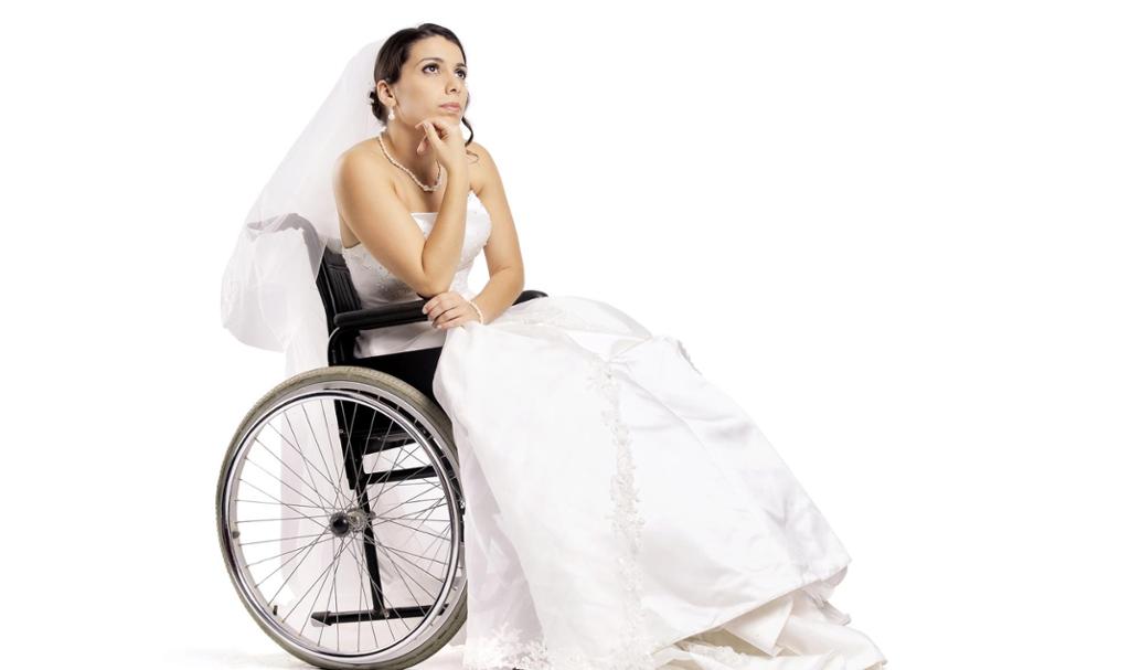 En brud i rullestol. Hun har hvit brudekjole, slør og pene smykker. Hun sitter og hviler haka på hånda og ser tankefullt opp i lufta. Foto.
