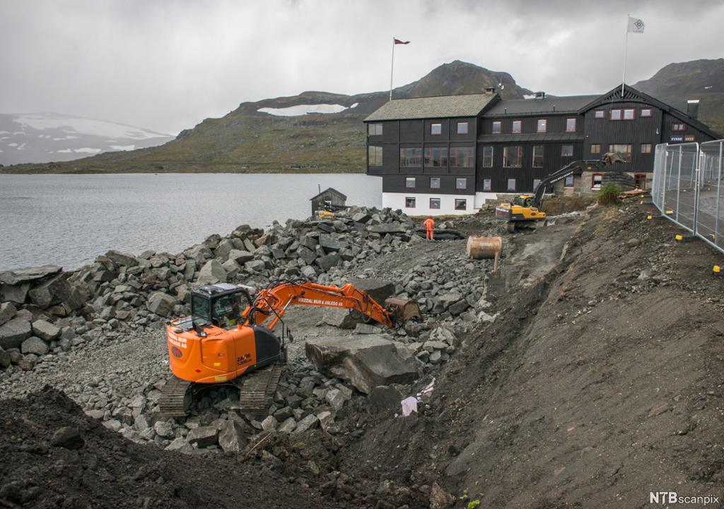 En oransje gravemaskin er i full sving på et anleggsfelt. I bakgrunnen ser vi et hotell, et vann og fjell. Foto.