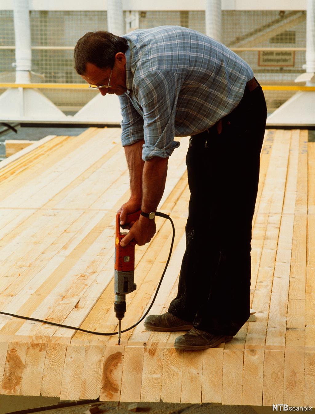 En mann bruker en elektrisk drill til å bore i planker. Foto.