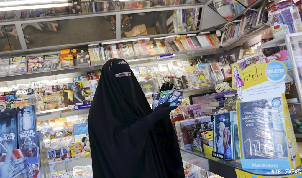 Kvinne med nikab og svart heildekkjande drakt i ein kiosk med magasin og CD-plater. Foto.