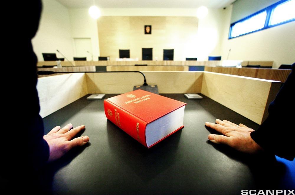 Et eksemplar av Norges lover ligger på en talerstol i en rettssal. Ved siden av den røde boka står to personer i svarte dressjakker med hver sin hånd mot bordflaten. Foto.