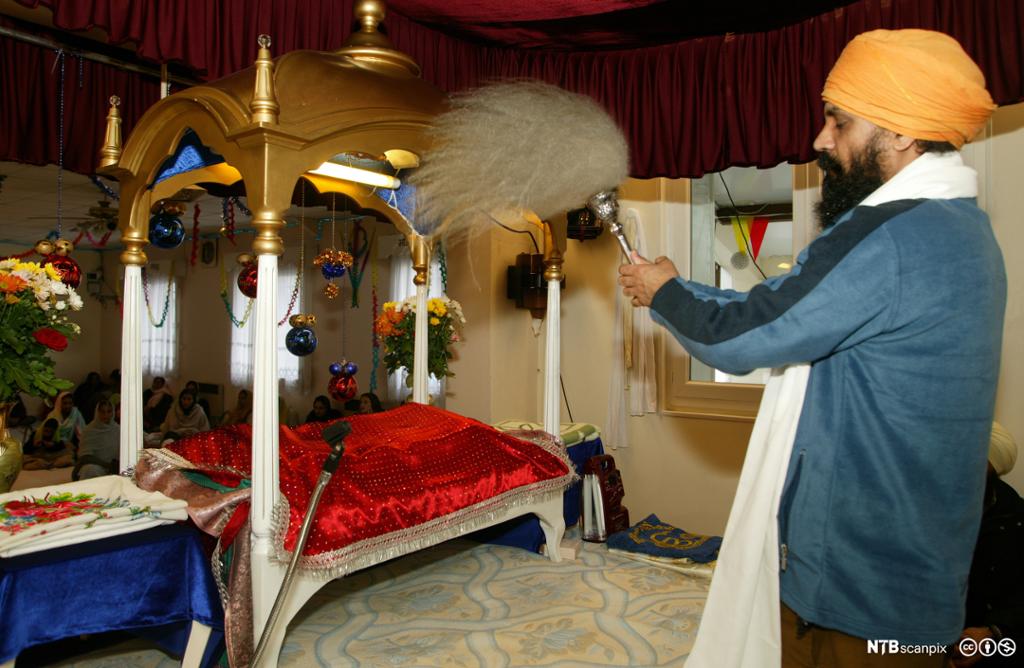 Mann med turban utfører eit rituale føre ei bok som ligg under eit raudt klede og ein baldakin. Foto.