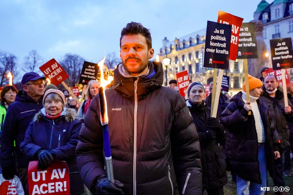 Rødt-leder Bjørnar Moxnes står med fakkel og løftet hode fremst i rekken av protestanter mot høye strømpriser. Protestantene bærer plakater der de krever politiske tiltak. Foto.