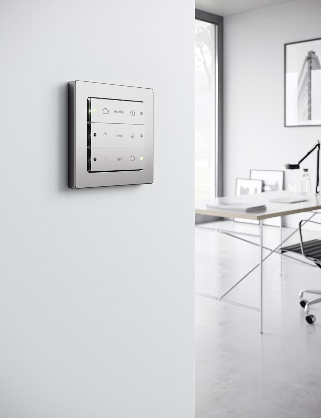 Vi ser en hvit vegg med brytere med symboler og LED-indikatorer. Bryterne kan styre oppvarming, persienner og belysning. I bakgrunnen ser vi en pult og en kontorstol. Foto.