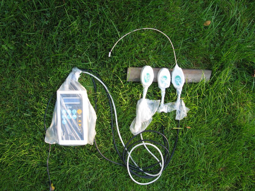 På graset ligg eit kvitt apparat pakka inn i ein plastpose. Ut frå posen går det kablar til tre mindre, kvite apparat som òg er pakka inn i plast kvar for seg. Foto