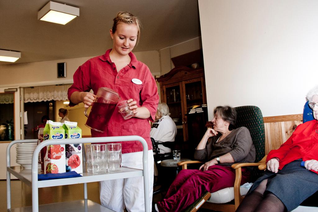 En kvinne serverer eldre på sykehjem drikke fra ei tralle. Foto.
