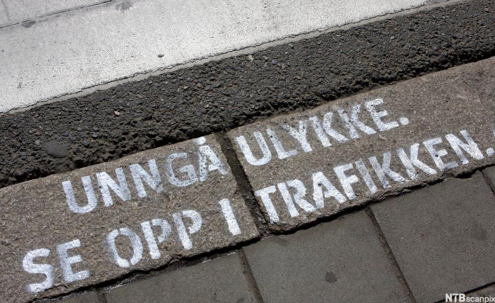 Fortauskant med følgjande tekst måla med kvite bokstavar på brustein: "Unngå ulykker. Se opp i trafikken." Foto.