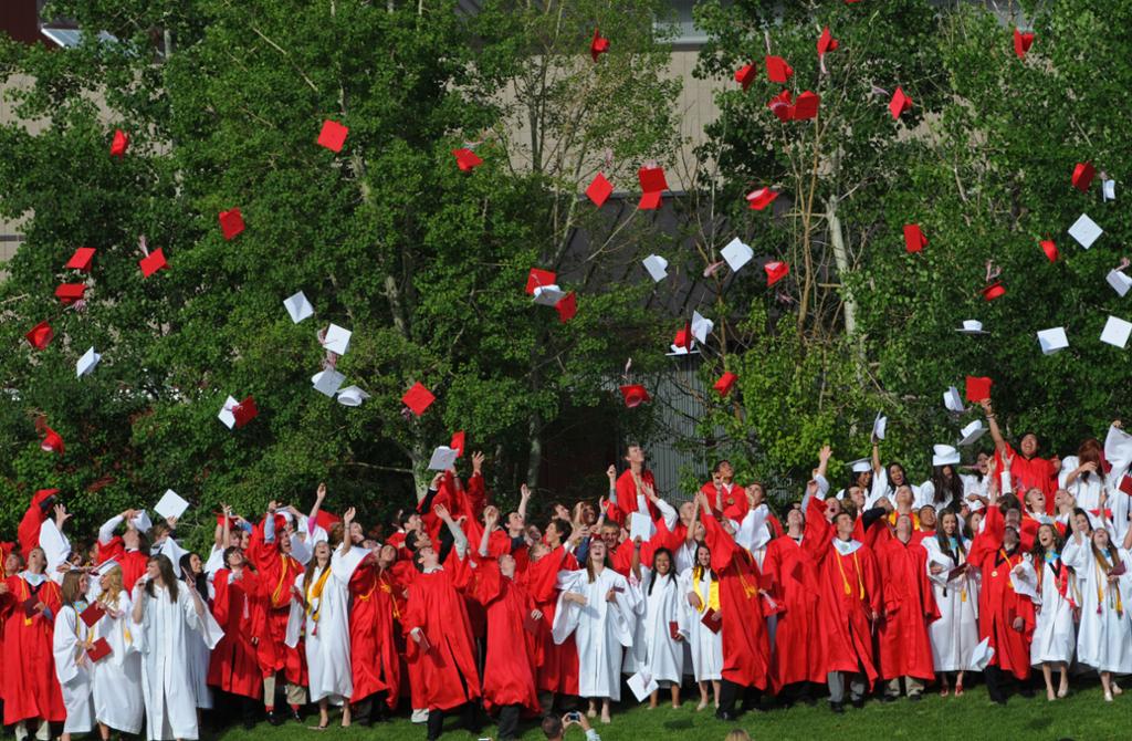 High school students dressed in red and white gowns throw their graduation caps in the air. 