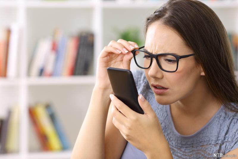 Kvinne med briller som myser og sliter med å lese innholdet på en mobiltelefon. Foto