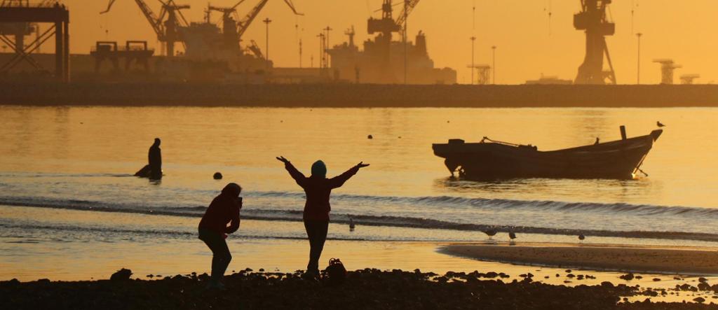 Personer og fugler på ei strand i solnedgang. Silhuettene til mange kraner på ei havn i bakgrunnen. Foto.