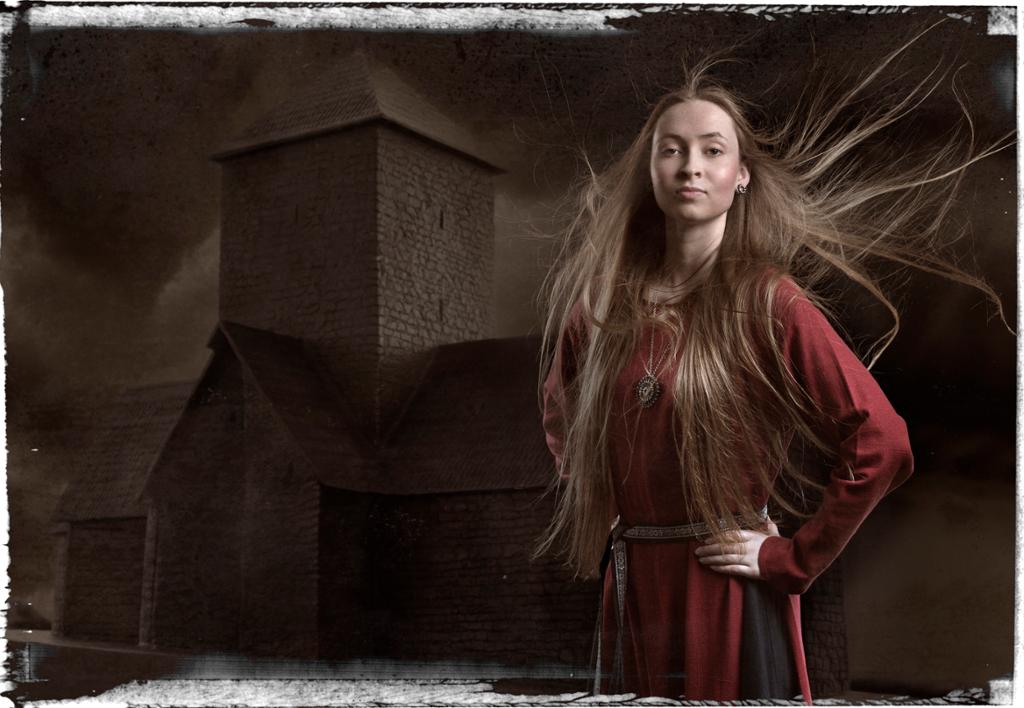 Ung kvinne med mørk rød kjole foran en steinkirke med et høyt tårn. Kvinnens lange hår flagrer i vinden, hun holden hendene på hoftene og ser bestemt foran seg. Manipulert foto.