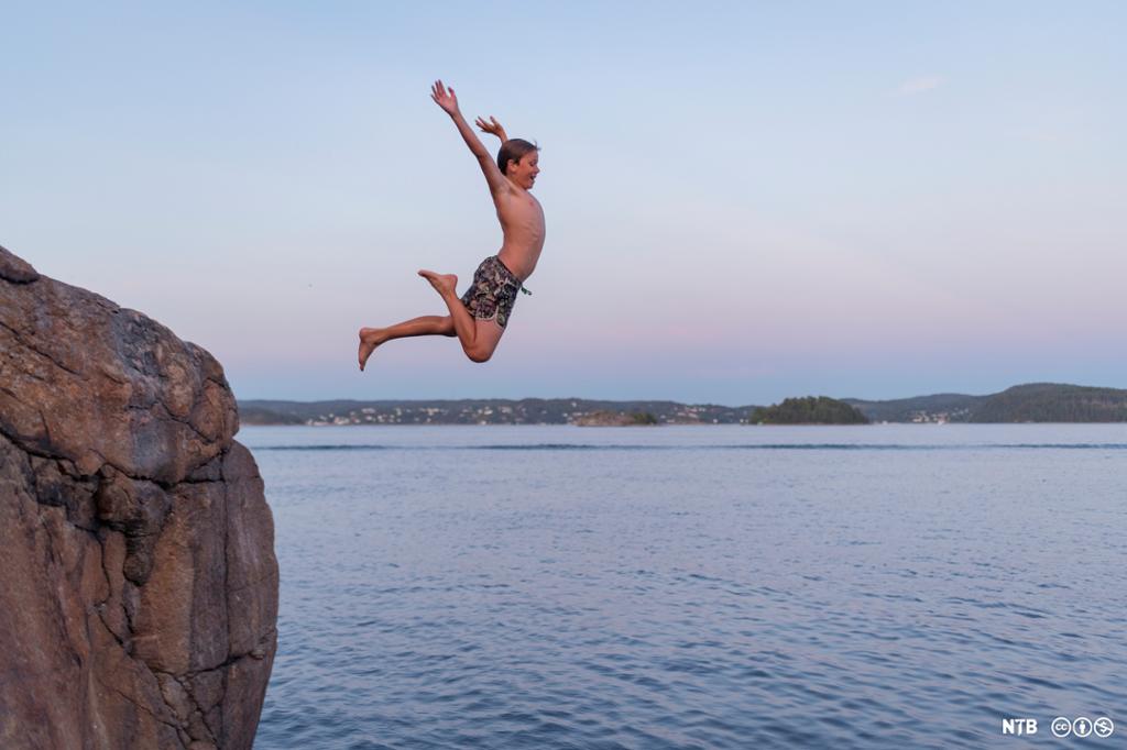 En gutt hopper fra en klippe og ned i vannet. Han smiler og holder armene høyt opp. Foto. 