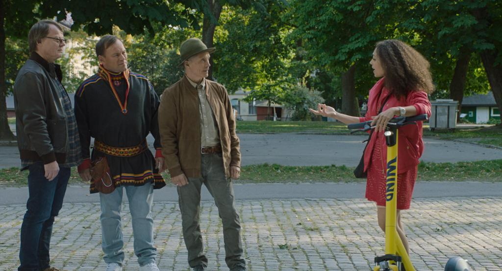 Utsnitt fra filmen "Koftepolitiet". Tre menn, den midterste med samekofte, står og snakker med ei urban dame med elektrisk sparkesykkel. Foto.