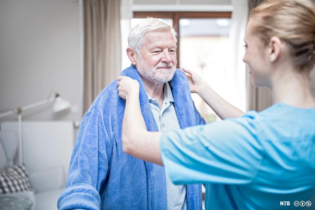 Helsefagarbeidar hjelper ein eldre mann med å kle på seg. Foto.