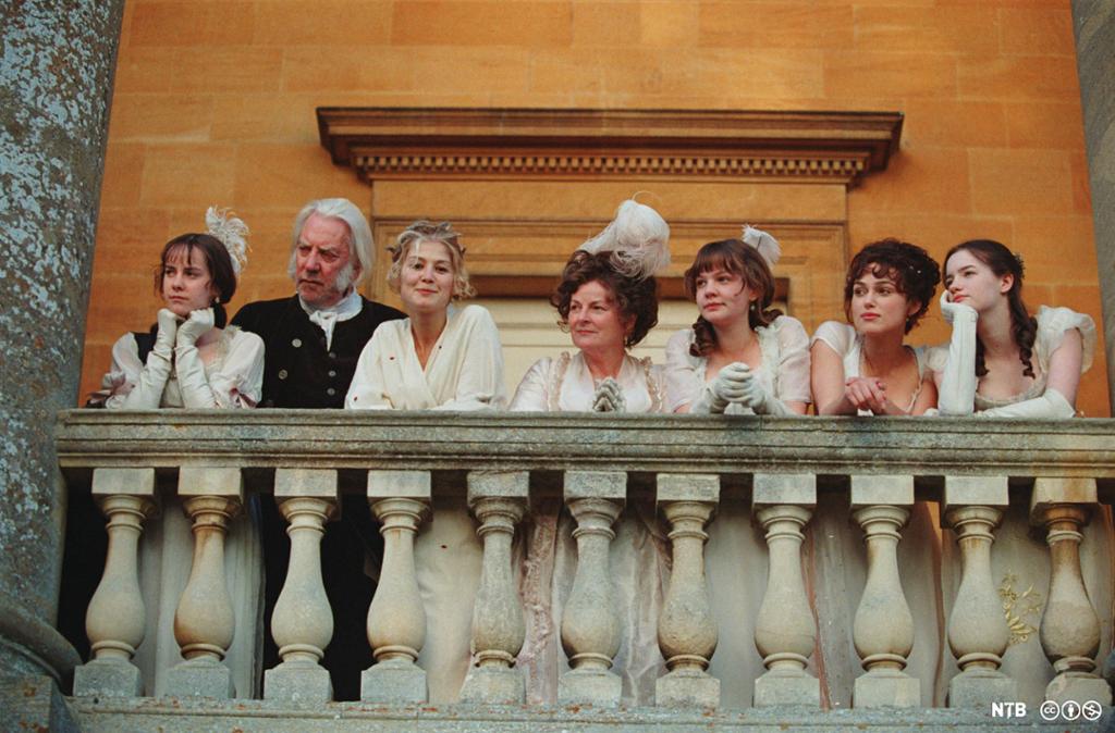 Still from the film Pride and Prejudice from 2005. We see six women and one man on a balcony. The women are dressed in white. The man wears a dark suit. 