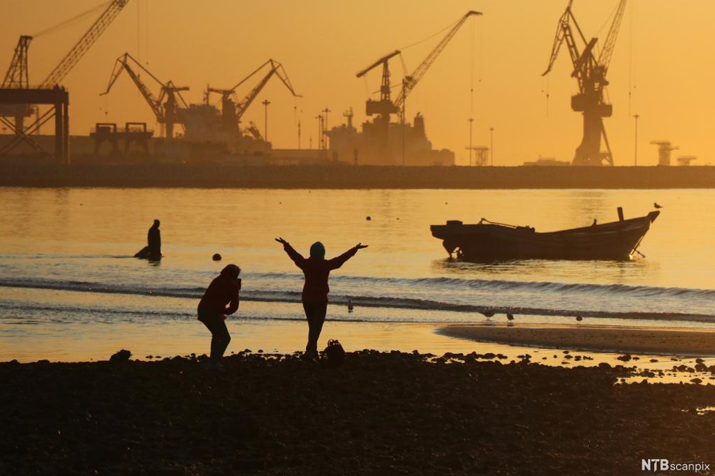 To personer står på ei strand i solnedgang. En liten båt ligger ute på sjøen, og svære heisekraner kan ses i bakgrunnen. Foto.