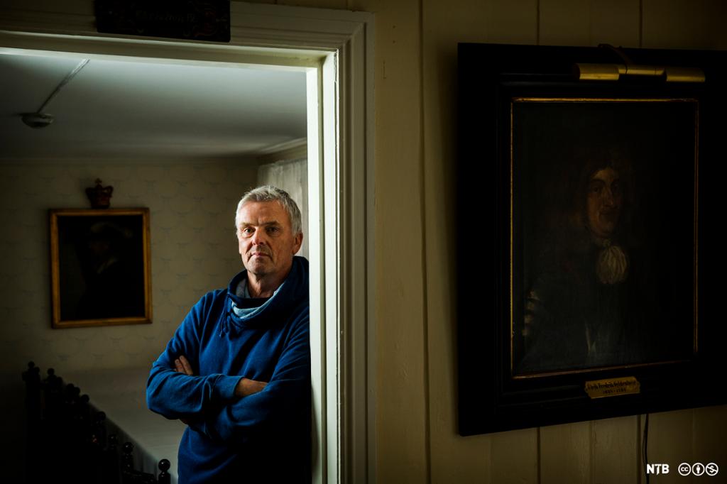 Programleder for Åndenes makt Tom Strømnæss står lent mot dørkarmen i åpningen mellom to rom i et eldre hus. I forgrunnen til høyre henger et maleri av en mann i 1700-tallsdrakt. Et annet maleri henger i bakgrunnen til venstre. Foto.