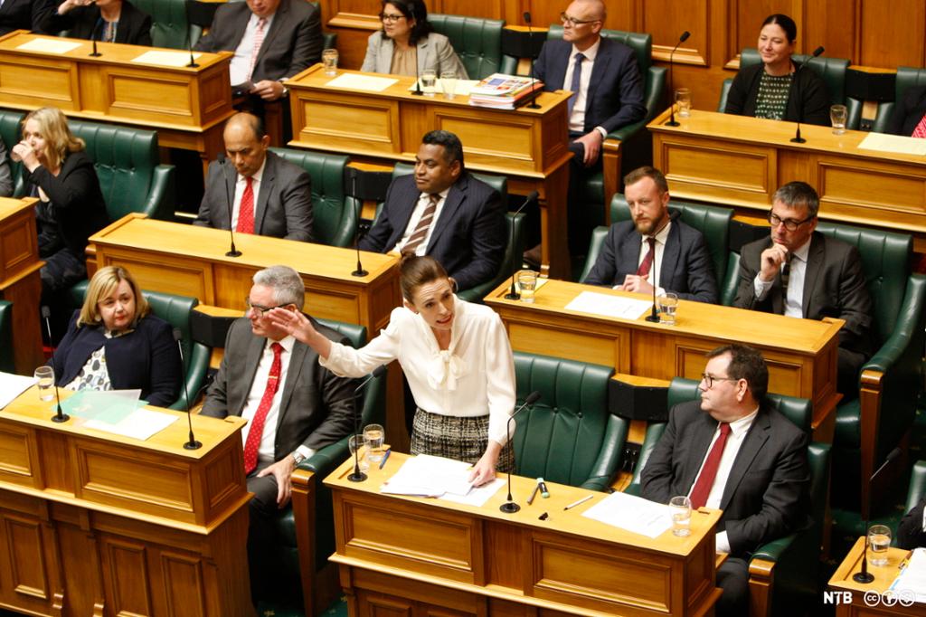 A room with many wooden desks. There are people in formal clothes seated at the desks. A woman dressed in a white blouse is standing, her left arm is raised in gesticulation. She is Prime Minister Jacinda Ardern. Photo. 