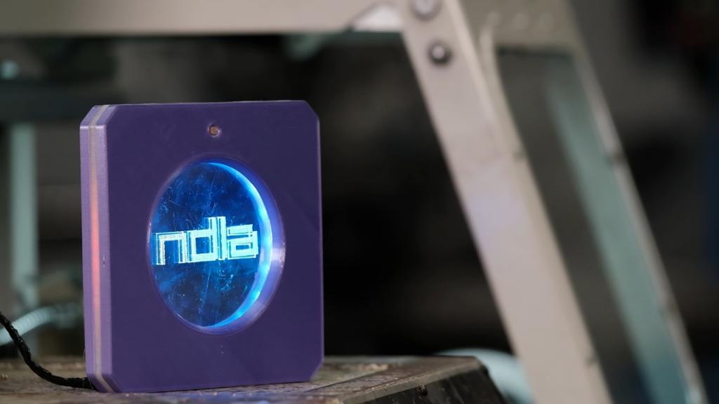 Et lite, firkanta skilt i lilla plast som står på et arbeidsbord. Midt i firkanten er det lagt inn en sirkel av blått pleksiglass med lysende NDLA-logo. Foto.