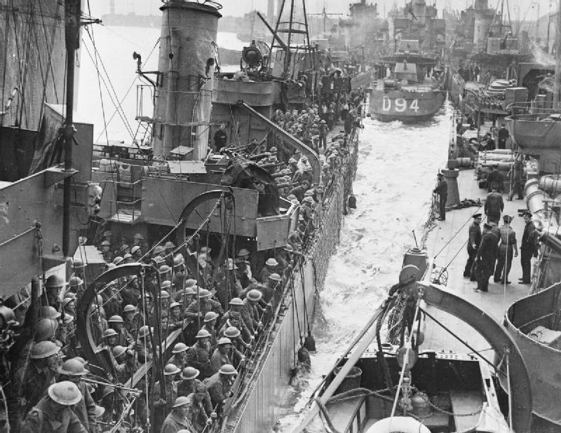 We see a ship full of soldiers wearing helmets.  Beside it, there is another ship where there are just a few people on deck. In the background ,there are more ships. Behind all the ships, we can vaguely see some buildings. The photo is in black and white. Photo.