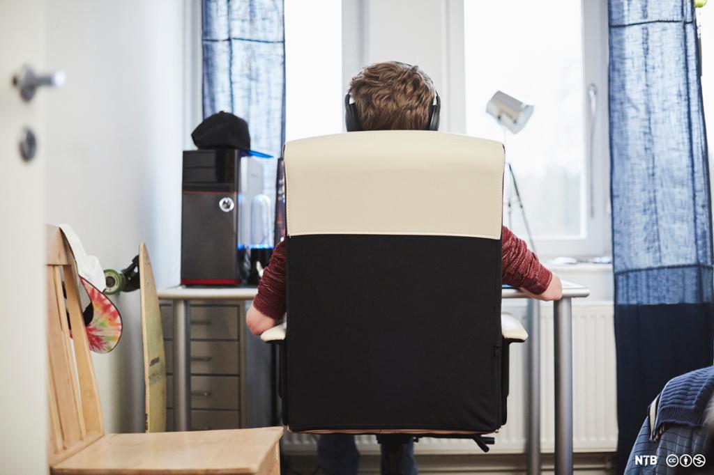 Person med hovudtelefonar sett bakfrå mens han sit i ein kontorstol framfor ein arbeidspult. Foto. 