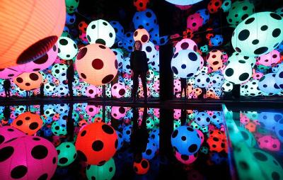 En person står inne i et rom med fargerike kuler som er dekorert med svarte sirkler. Veggene, gulvet og taket er dekket av speil, slik at elementene gjentas. Foto.