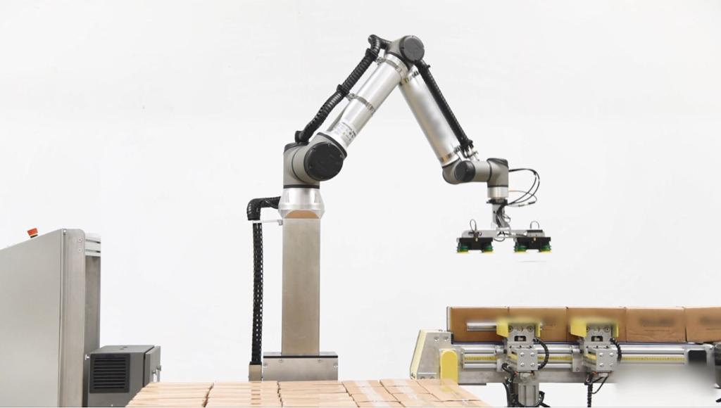 Robot med sugekoppverktøy som står ved et samleband med pappesker. Ved sida av samlebandet står det en pall med stabla pappesker. Foto.