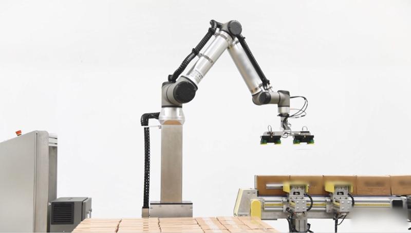 Robot med sugekoppverktøy som står ved et samlebånd med pappesker og en palle med stablede pappesker. Foto