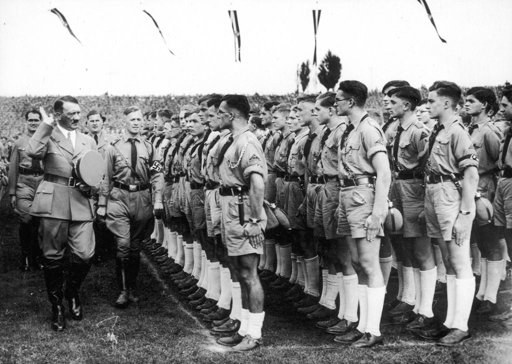 Hitler går foran rekkene av oppstilte unge menn i Hitlerjugend-uniformer. De har knebukser, kortermede skjorter og hvite strømper. Foto.