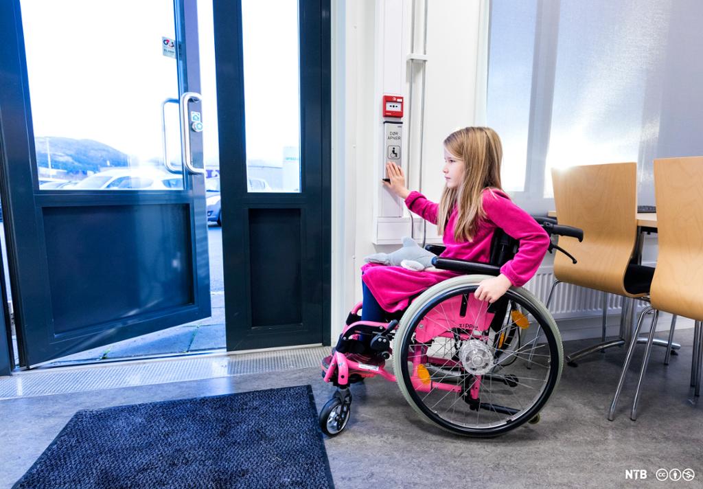 Ung jente i rosa rullestol åpner ei dør ved hjelp av automatisk døråpner. Foto.