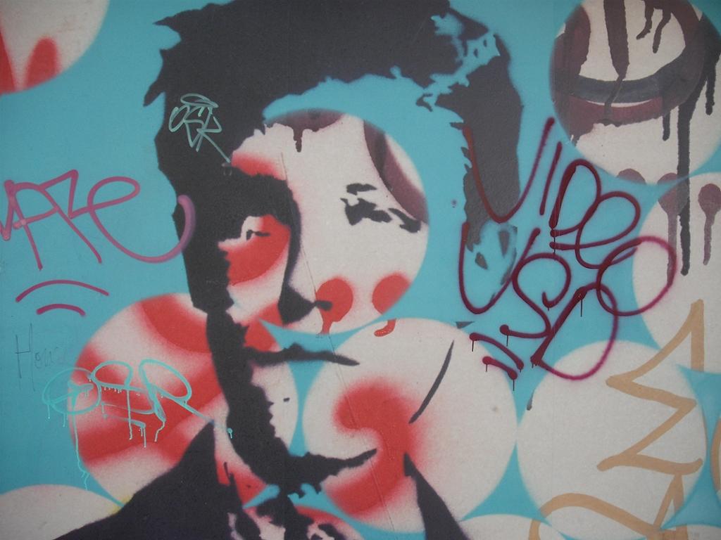 En vegg som er spraymalt med graffiti. Midt mellom all graffitien er det plassert et sjablongtrykk av portrettet av en ung mann. Mannen har et  smalt ansikt og kort, mørkt hår. Foto.