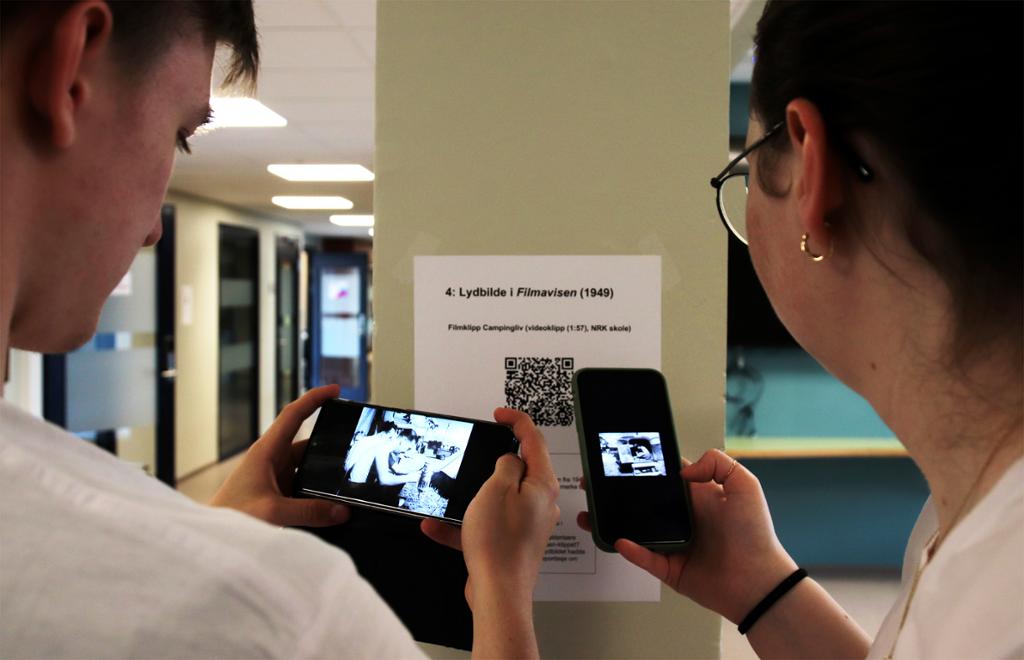 To elever ser på et videoklipp i svart-hvitt på mobiltelefonene sine. Et ark med en QR-kode er teipet på en søyle foran dem. Foto.