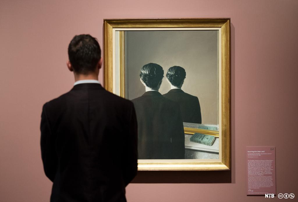 Photo: We see a dark-haired man in a suit standing with his back to the camera looking at the painting of the back of a dark-haired man in a suit looking at the back of a dark-haired man in a suit. (A reflection, or a painting within the painting?)