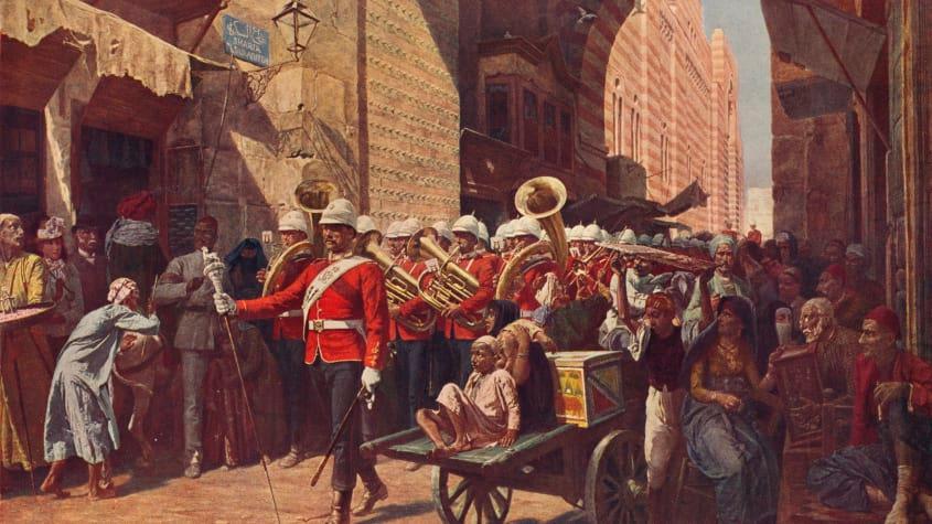 A British marching band dressed in red uniforms is walking through the narrow roads of a town in Egypt. The native people are watching: they have serious expressions on their faces. 
