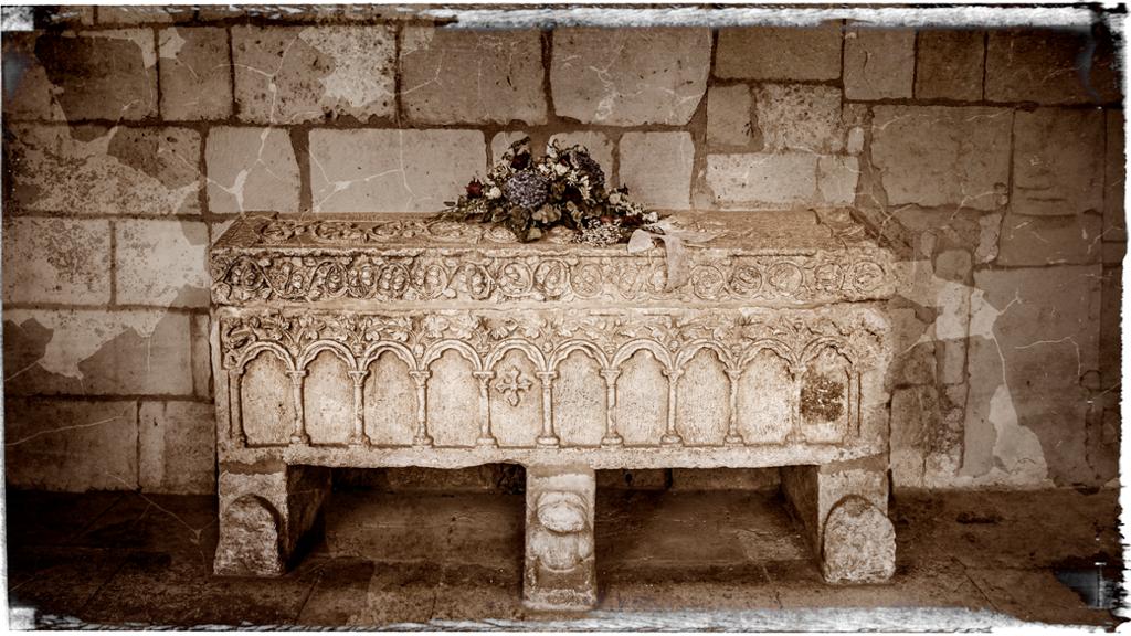 En steinsarkofag inntil en murvegg. Sarkofagen er dekorert med inngraverte mønster og det ligger blomster på lokket. Manipulert foto.