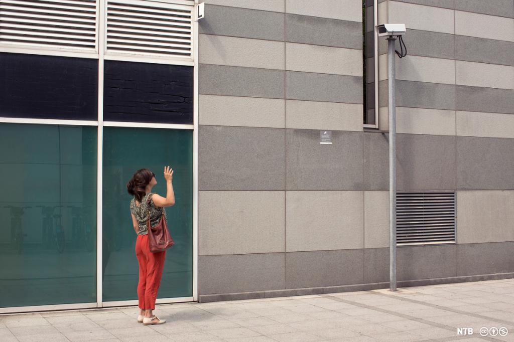 Kvinne står på et fortau og vinker til et overvåkingskamera. Foto.
