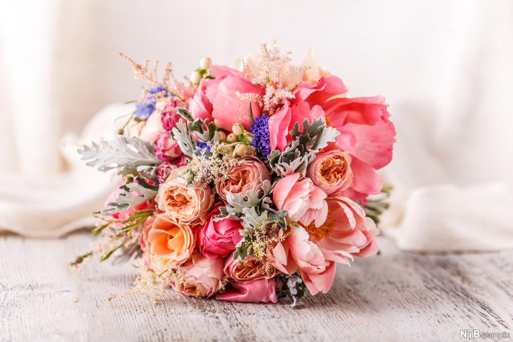 Fotografi av en blomsterbukett bestående av roser og peoner i ulike rosatoner. Buketten ligger på ei slitt, lysfarga bordplate av tre.