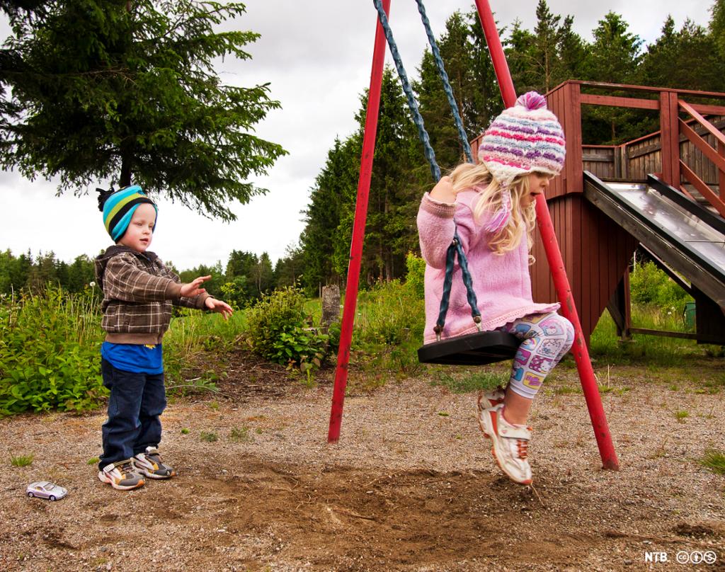 Gutt på cirka tre år dytter huska til en jente på en lekeplass ved skogen. Foto. 